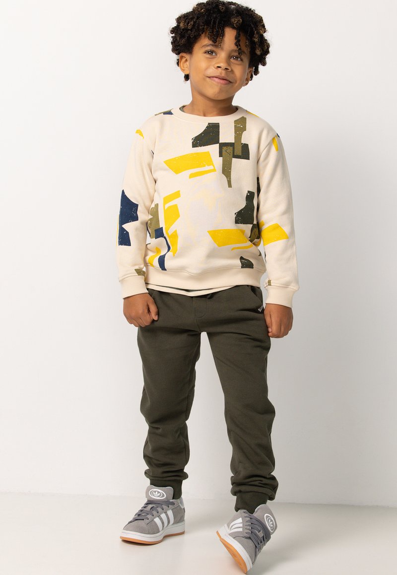 Beige sweatshirt met abstracte gele, groene en blauwe patronen; gecombineerd met olijfgroene sweatpants en grijze sneakers met witte strepen.