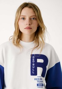 Felpa in fleece caratterizzata da un corpo bianco con maniche blu, grande patch blu con la lettera "R" e testo "RED BULL TEAM" in blu e bianco.
