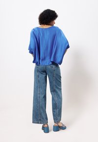 Blaue, fließende Bluse mit weiten Ärmeln, kombiniert mit hoch sitzenden, hellblauen, weit geschnittenen Jeans und blauen Blockabsatzsandalen.