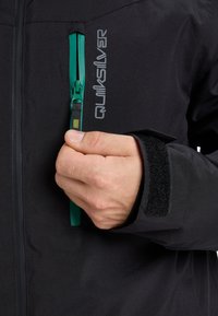 Schwarze Jacke mit grünem Reißverschluss, mit einem Logo und Klettverschluss an den Manschetten. Das Material wirkt glatt und wetterfest.