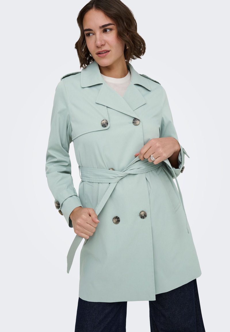 Femme ajustant une ceinture sur un trench-coat vert clair à double boutonnage avec de gros boutons, debout devant un fond blanc uni.