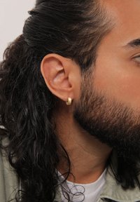 Cercel de aur în formă de cerc pe urechea unui model, având un finisaj neted și lucios, cu o formă rotunjită, complementând părul închis și creț al modelului.