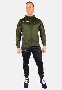 Felpa con zip verde scuro e cordino, abbinata a pantaloni da jogging neri. Logo bianco sulla felpa e sui pantaloni, indossata con sneakers nere e bianche.