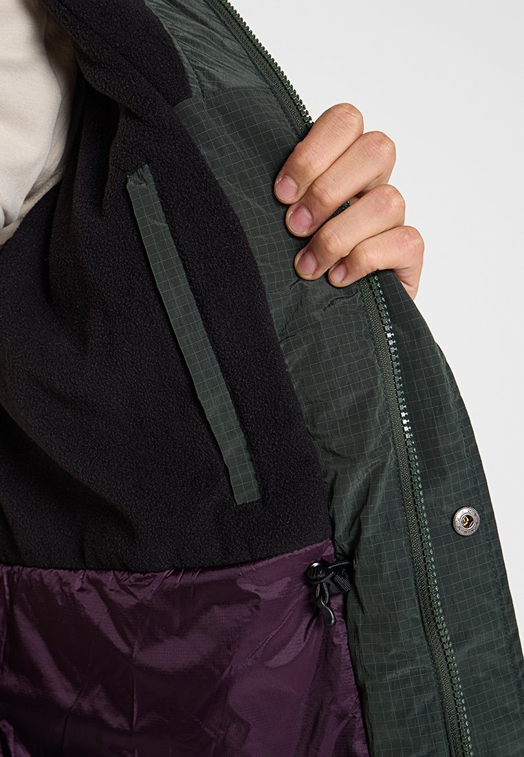 Ręka otwierająca ciemnozieloną kurtkę, odsłaniająca wewnętrzną czarną polarową podszewkę i fioletowy pikowany materiał z zapięciem na zamek i guziki snap.