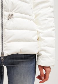 Bílá péřová bunda s prošívanými částmi, stříbrným zipem a ochranným létem. Tkanina vypadá hladce a leskle. Nosit s modrými džíny.