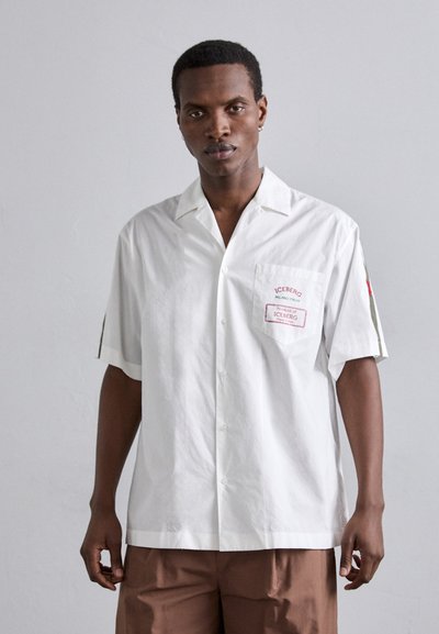 Chemise blanche à manches courtes avec col ouvert, dotée d'une poche poitrine avec un texte rouge, et d'une coupe décontractée. Le matériau semble léger.