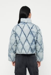 Vrouw met lang zwart haar, gekleed in een lichtblauwe gewatteerde jas met ruitpatronen, gecombineerd met donkergrijze cargobroek, staand met haar rug naar de kijker gekeerd.