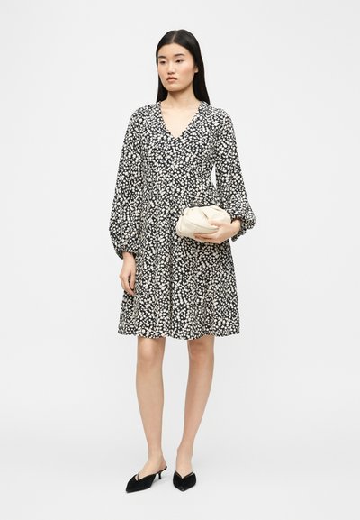 Γυναίκα που φοράει μακρυμάνικο φόρεμα με ασπρόμαυρα λουλούδια, κρατώντας ένα μικρό λευκό φάκελο και φορώντας μαύρες μυτερές γόβες, στέκεται μπροστά σε άσπρο φόντο.