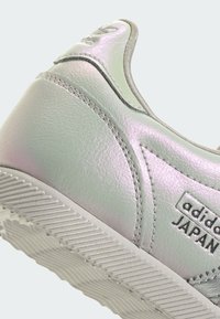 Sudraba krāsas spīdīgs ādas sporta apavu pāris ar teksturētu gumijas zoli, izšūtu dizainu un uz sāniem iespiestu zīmolu.