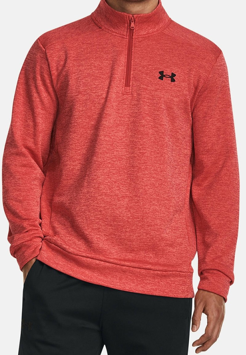 Homme portant un sweat-shirt à col quart-zip rouge avec un logo Under Armour noir sur la poitrine gauche et un pantalon noir.
