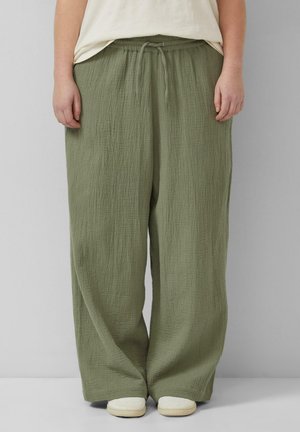 Person trägt locker sitzende olivgrüne, strukturierte Hosen mit Kordelzug in der Taille und weißen Freizeitschuhen und steht vor einem schlichten Hintergrund.