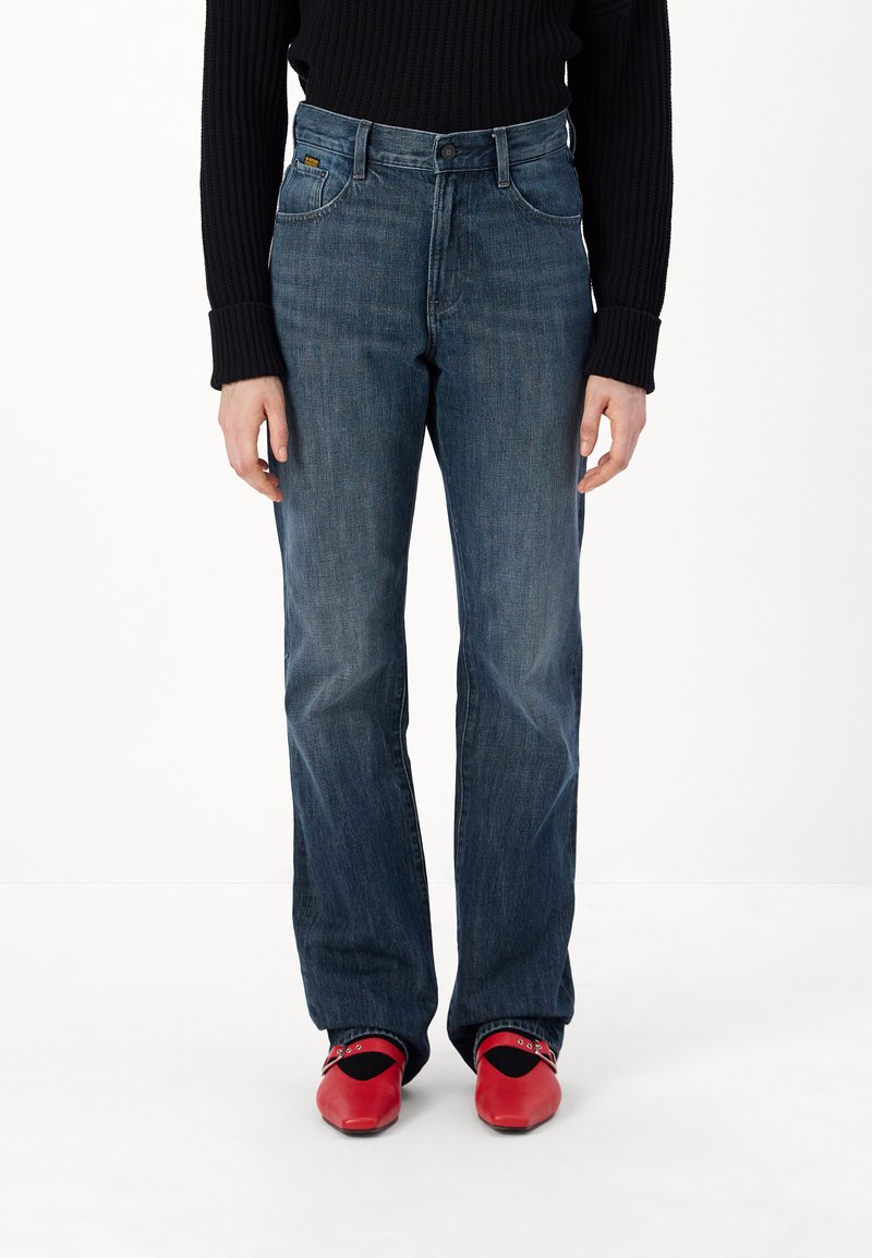 Blue denim flared jeans with a medium wash, featuring a classic five-pocket design and a zip fly, worn with red clogs.