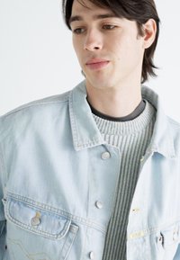Chaqueta de denim azul claro con un corte clásico, que presenta dos bolsillos en el pecho, botones de presión plateados y detalles de costura sutiles. Lleva puesta una sudadera de punto gris por debajo.