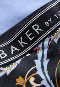Primo piano di un tessuto con un elastico nero etichettato "BAKER" e un motivo floreale nei toni di blu, verde e oro sul tessuto.