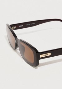 Čierne obdĺžnikové slnečné okuliare s hnedými šošovkami. Vyznačujú sa minimalistickým dizajnom a zlatým detailom na boku s názvom značky. Hladký povrch.