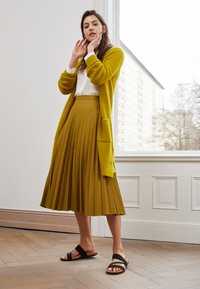Cardigan en tricot jaune moutarde, jupe plissée en vert olive, et sandales noires. Design simple, tissu texturé et style superposé.