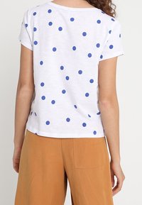 Camiseta blanca con lunares azules, de mangas cortas y cuello redondo. Combinada con pantalones anchos color beige. La tela parece suave y ligera.