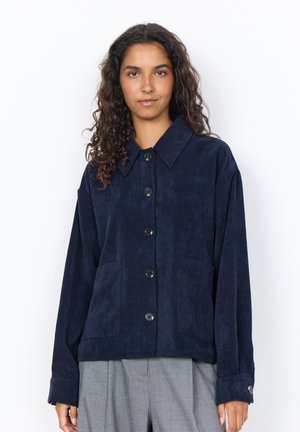 Woman with curly hair wearing a navy blue corduroy button-up jacket and gray pants, standing against a white background.