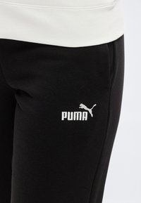 Μαύρο παντελόνι φόρμας με πλαϊνές τσέπες και λευκό λογότυπο Puma στο μηρό, φορεμένο με λευκό τοπ.