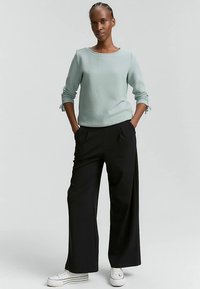 Világoszöld texturált pulóver háromnegyedes ujjakkal és oldalsó megkötőkkel, laza fekete nadrággal párosítva. A ruhát fehér tornacipő egészíti ki.