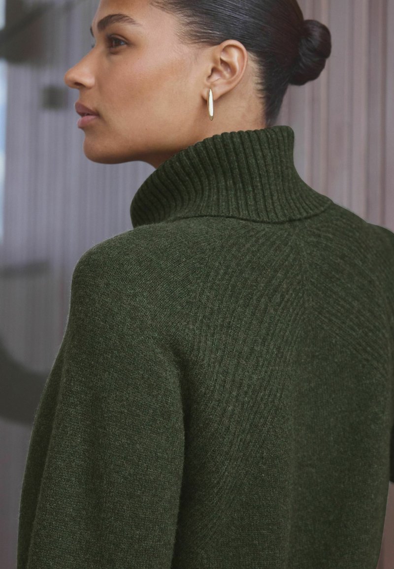 Grüner Strickpullover mit hohem geripptem Kragen und strukturiertem Muster. Das Modell trägt ein Paar schlichte goldene Creolen.