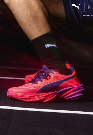 Tek tekačev z rdečo tekaško čeveljiko Puma Nitro s črnim logotipom Puma in črno nogavico na štartni črti steze.