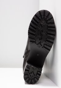 Suela de bota negra con surcos profundos y patrones en forma de cruz, que muestra una talla 37, colocada en posición vertical contra paredes blancas de esquina.