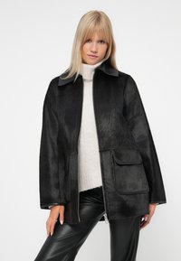 Giacca in pelliccia sintetica nera con zip anteriore, grandi tasche a toppa e colletto in pelle nera. Indossata sopra un maglione chiaro e pantaloni neri.