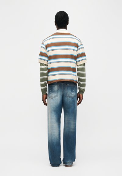 Ενήλικας που φοράει ξεθωριασμένα μπλε τζιν και στρωμμένα ριγέ πουκάμισα σε καφέ, μπλε, λευκές και πράσινες αποχρώσεις, στέκεται με την πλάτη γυρισμένη προς ένα απλό φόντο.