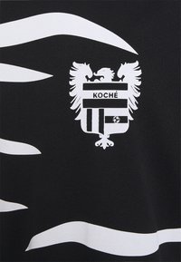 Schwarzer Stoff mit weißen, wellenförmigen Streifen und einem zentralen Emblem mit einem Doppeladler sowie horizontalen Balken, die "KOCHÉ" beschriftet sind.