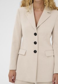 Blazer beige sur mesure avec une texture lisse, un col à revers cranté et quatre boutons noirs. Il comprend des poches latérales et une silhouette ajustée.