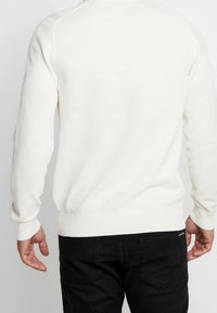 Maglione bianco a maglia con tessuto testurizzato, polsini e vita a coste, mostrato di schiena con jeans neri visibili in vita.