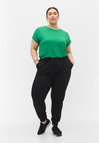 T-shirt verde a maniche corte abbinato a pantaloni jogger neri con cintura elastica e tasche laterali, completato da scarpe sportive nere.