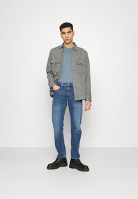 Chemise grise à boutons sur un t-shirt bleu, associée à un jean bleu et des bottes noires. La chemise a deux poches poitrine et une texture douce.