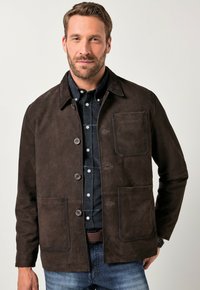 Giacca in suede marrone con colletto a camicia, chiusura frontale a bottoni, due grandi tasche e una finitura texturizzata. Indossata sopra una camicia a quadri blu.
