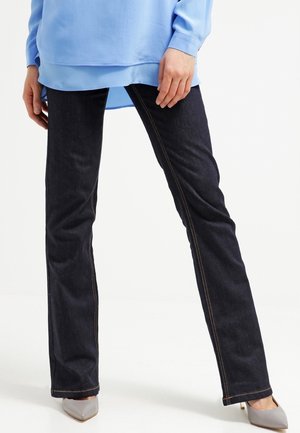 Persoon die donkerblauwe rechtvallende jeans, een lichtblauw lang overhemd en grijze pumps met puntige neus draagt, staand met de benen iets uit elkaar.