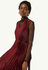 Rotes plissiertes Kleid mit V-Ausschnitt, über einem transparenten schwarzen Mesh-Longsleeve-Shirt getragen. Hat eine taillierte Taille und einen eleganten Fall.