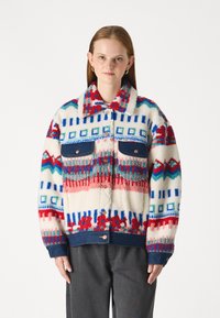 Chaqueta de forro polar con un patrón geom�étrico multicolor en rojo, azul y crema, con acentos de mezclilla y dos bolsillos frontales.