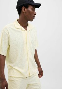 Camisa amarilla de manga corta con un patrón de olas texturizado, con botones al frente y cuello abierto. La tela parece ligera con un acabado suave.
