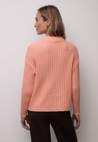 Pull en tricot rose avec une texture côtelée, col montant et manches longues, associé à un pantalon marron foncé. Design simple et moderne.
