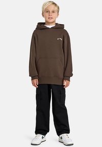 Blondé dítě stojící, oblečené v hnědé mikině s kapsou, černých cargo kalhotách a bílých teniskách se zeleným logem.