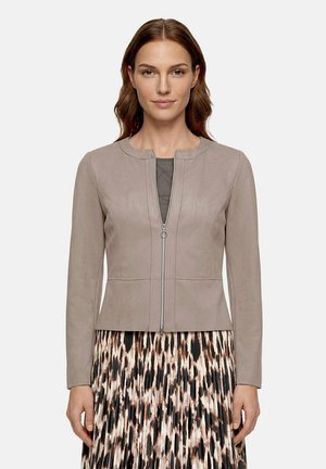 Femme portant une veste taupe zippée sur une jupe plissée à motifs, debout devant un fond clair uni.