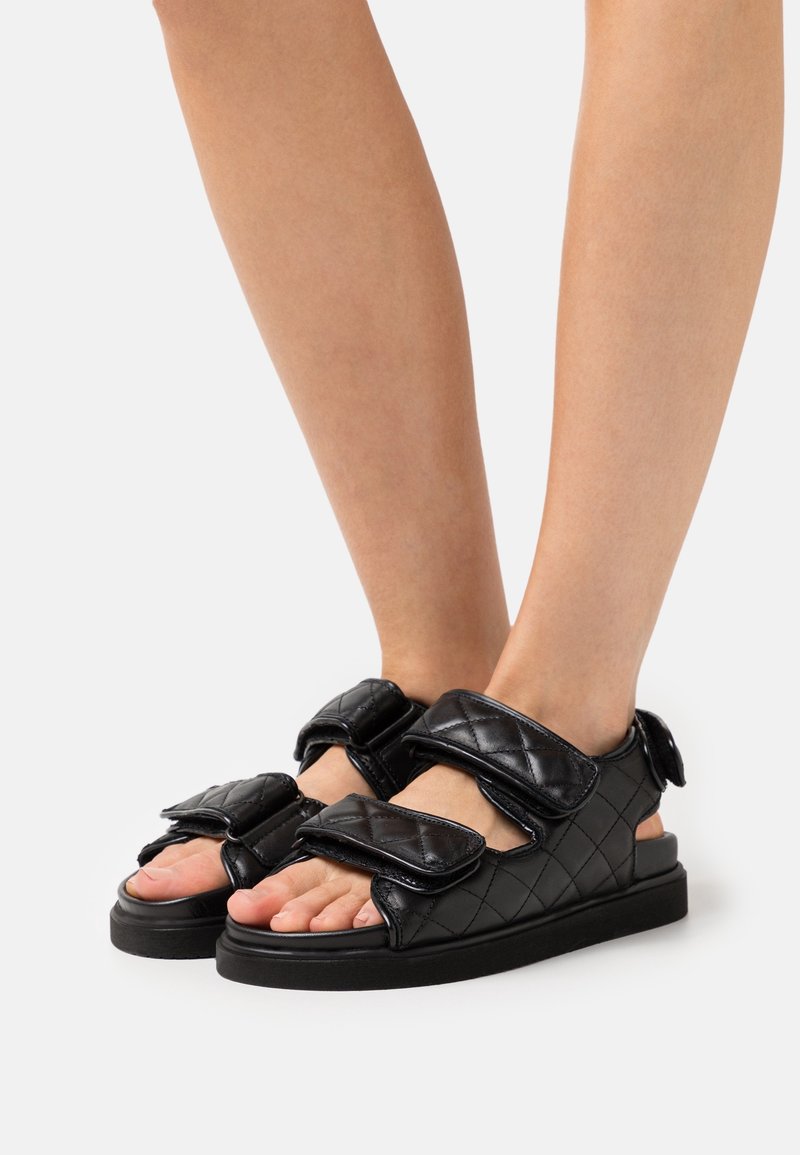 Sandales noires matelassées avec deux larges bandes Velcro, semelle plate en caoutchouc, design à bout ouvert et texture lisse.