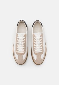 Baskets en daim blanc et beige avec un accent talon noir. Dotées de lacets blancs plats et d'une semelle en caoutchouc texturée. Marquage à l'intérieur de la chaussure.