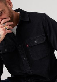 Camicia di velluto a coste nera con due tasche frontali, chiusura con bottoni e superficie texturizzata. Presenta un piccolo etichetta rossa sulla tasca sinistra.