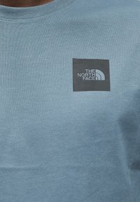 Ανοιχτό μπλε βαμβακερό μπλουζάκι με τετράγωνο μαύρο λογότυπο που φέρει το κείμενο "THE NORTH FACE" και κυρτό σχέδιο, τοποθετημένο στο πάνω αριστερό μέρος του στήθους.