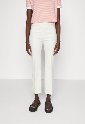 Persona con pantalones blancos recortados, camiseta de manga corta a rayas rosas y blancas, y sandalias negras con tiras, de pie contra un fondo liso.