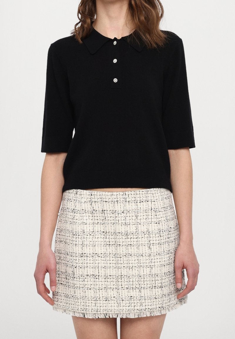 Femme portant un haut noir à manches courtes avec col et trois boutons, ainsi qu'une mini-jupe taille haute en tweed blanc avec des fils noirs.