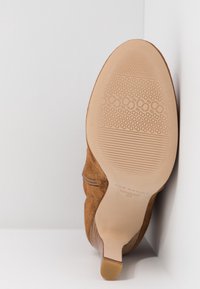 Chaussure à talon haut en daim marron vue de la semelle, avec des motifs antidérapants texturés et un design pointu à l'avant.