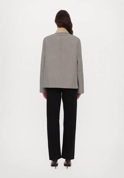 Houndstooth mintázatú dzseki bézs gallérral, fekete egyenes szárú nadrág felett viselve, hátulról nézve, hosszú ujjakkal és laza szabással.